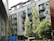 A modern, multi-story residential building with large glass balconies surrounded by lush green trees. The building has a sleek design with a mix of metal and glass surfaces. In the foreground, there is a blue dumpster and several trees providing some foliage.