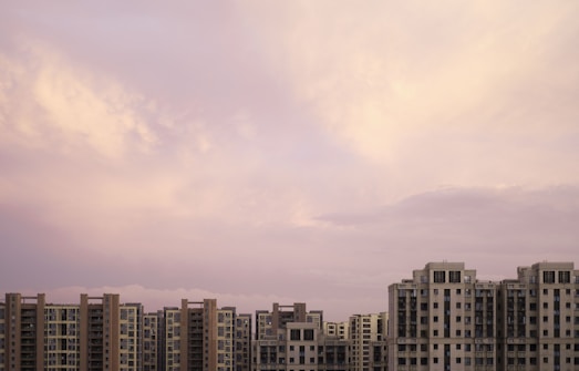 A series of modern high-rise apartment buildings stretch across the skyline under a soft, pastel-colored sky. The buildings feature a geometric arrangement of windows and balconies, and the sky is filled with light shades of pink and purple clouds.