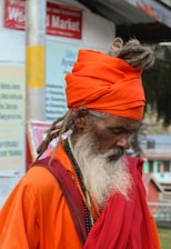 An elderly person with a long white beard is wearing an orange turban and robes, standing in a seemingly urban setting. Their hair is styled in dreadlocks, and they appear deep in thought or meditation. The clothing includes beaded necklaces and layered fabric. The background shows signs and blurry buildings, indicating a public area.