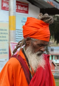 An elderly person with a long white beard is wearing an orange turban and robes, standing in a seemingly urban setting. Their hair is styled in dreadlocks, and they appear deep in thought or meditation. The clothing includes beaded necklaces and layered fabric. The background shows signs and blurry buildings, indicating a public area.