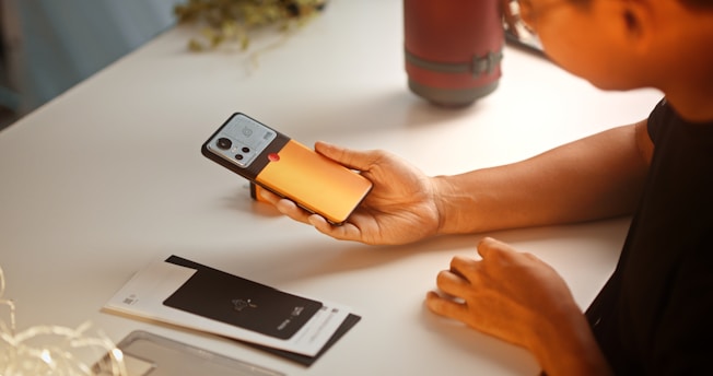 A person holding a smartphone with an orange and black case in their hand. The phone is positioned over a white desk with a notebook or pamphlet visible nearby. The person is wearing a short-sleeved shirt, and there is a cylindrical red object in the background.