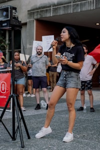 A person wearing a black t-shirt and denim shorts is speaking into a microphone during an outdoor event. They are holding a phone in one hand and are standing near a speaker. A group of people in the background hold signs indicating a protest or rally setting.