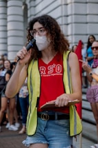 A person with curly hair is speaking into a microphone, wearing a face mask, glasses, a red shirt with 'RSO' printed on it, a yellow reflective vest, and denim shorts. They are holding a clipboard and appear to be part of a gathering or rally. In the background, a crowd of people is visible, some of them holding signs or banners.
