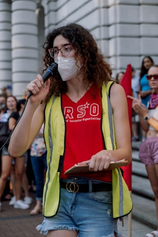 A person with curly hair is speaking into a microphone, wearing a face mask, glasses, a red shirt with 'RSO' printed on it, a yellow reflective vest, and denim shorts. They are holding a clipboard and appear to be part of a gathering or rally. In the background, a crowd of people is visible, some of them holding signs or banners.