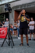 A person is speaking into a microphone in front of a crowd. They are holding a plastic water bottle and wearing a black shirt with a large yellow smiley face. In the background, several people are gathered, some with camera equipment, suggesting a possible public demonstration or gathering. A red banner on the left includes partially visible text.