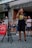 A person is speaking into a microphone in front of a crowd. They are holding a plastic water bottle and wearing a black shirt with a large yellow smiley face. In the background, several people are gathered, some with camera equipment, suggesting a possible public demonstration or gathering. A red banner on the left includes partially visible text.