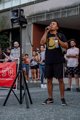 A person is speaking into a microphone in front of a crowd. They are holding a plastic water bottle and wearing a black shirt with a large yellow smiley face. In the background, several people are gathered, some with camera equipment, suggesting a possible public demonstration or gathering. A red banner on the left includes partially visible text.