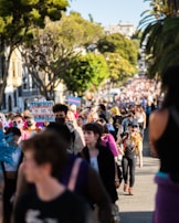 A large crowd of people is gathered outdoors, with many individuals walking along a street lined with trees. The focal group is holding signs, one of which reads 'Trans Rights Are Human Rights.' The crowd is diverse, and some people are wearing masks and vibrant clothing. The atmosphere is lively, and the surrounding environment includes trees and buildings.