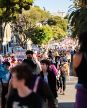 A large crowd of people is gathered outdoors, with many individuals walking along a street lined with trees. The focal group is holding signs, one of which reads 'Trans Rights Are Human Rights.' The crowd is diverse, and some people are wearing masks and vibrant clothing. The atmosphere is lively, and the surrounding environment includes trees and buildings.