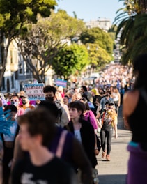 A large crowd of people is gathered outdoors, with many individuals walking along a street lined with trees. The focal group is holding signs, one of which reads 'Trans Rights Are Human Rights.' The crowd is diverse, and some people are wearing masks and vibrant clothing. The atmosphere is lively, and the surrounding environment includes trees and buildings.