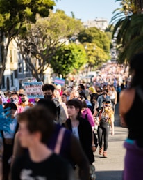 A large crowd of people is gathered outdoors, with many individuals walking along a street lined with trees. The focal group is holding signs, one of which reads 'Trans Rights Are Human Rights.' The crowd is diverse, and some people are wearing masks and vibrant clothing. The atmosphere is lively, and the surrounding environment includes trees and buildings.