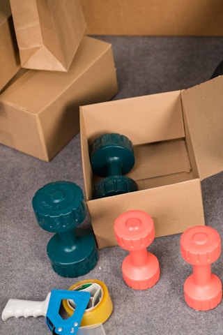 Several cardboard boxes and dumbbells are arranged on a carpeted floor. A pair of teal dumbbells is inside an open box, while another is positioned outside. Two coral-colored dumbbells are placed nearby. In the foreground, a blue and white tape dispenser and a roll of packing tape are visible.