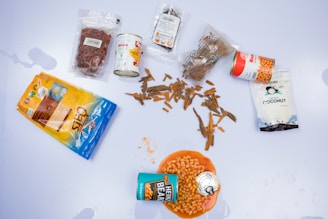 A collection of packaged food items, including dried chili peppers, canned food, and coconut chips, arranged on a white surface. Various packages are open, with one can of beans spilling its contents onto an orange bowl.