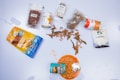 A collection of packaged food items, including dried chili peppers, canned food, and coconut chips, arranged on a white surface. Various packages are open, with one can of beans spilling its contents onto an orange bowl.