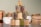 A pyramid of glass jars filled with various grains and legumes is carefully arranged on a dark surface. The jars contain diverse items such as beans, lentils, and rice, and are sealed with golden metal lids. In the background, there are blurred sacks of similar products, suggesting a pantry or storage setting.