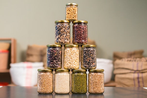 A pyramid of glass jars filled with various grains and legumes is carefully arranged on a dark surface. The jars contain diverse items such as beans, lentils, and rice, and are sealed with golden metal lids. In the background, there are blurred sacks of similar products, suggesting a pantry or storage setting.