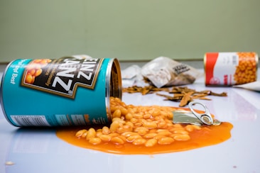 A can of Heinz baked beans is lying on its side with beans spilling out in orange sauce. There are other items in the background including a similar can of beans, some dried food items, and a blurred bag. The scene takes place on a light-colored countertop.