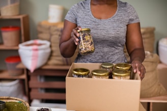 A person is holding a jar of mixed nuts or seeds and placing it in a cardboard box filled with similar jars. The setting appears to be a storage or packing area with shelves and bags in the background.