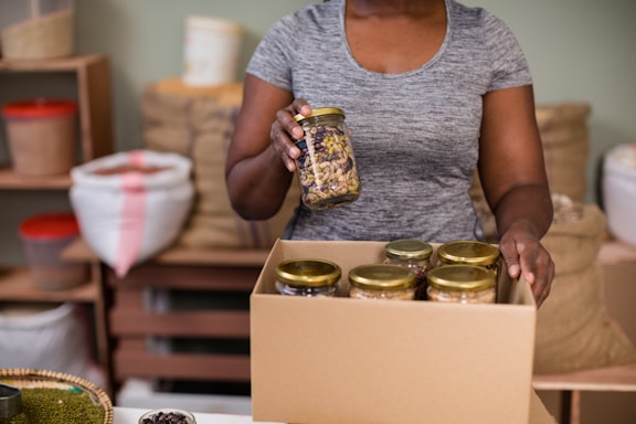 A person is holding a jar of mixed nuts or seeds and placing it in a cardboard box filled with similar jars. The setting appears to be a storage or packing area with shelves and bags in the background.
