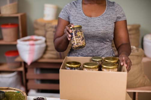 A person is holding a jar of mixed nuts or seeds and placing it in a cardboard box filled with similar jars. The setting appears to be a storage or packing area with shelves and bags in the background.