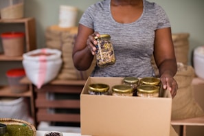 A person is holding a jar of mixed nuts or seeds and placing it in a cardboard box filled with similar jars. The setting appears to be a storage or packing area with shelves and bags in the background.