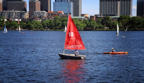 A sailboat with a prominent red sail and the number 18, featuring 'MIT' written on it, traverses a body of water with a person onboard. To the right, an orange kayak with a single individual paddling is visible. In the background, several other sailboats and the urban skyline of a city, marked by modern high-rise buildings and green trees, can be seen under a clear blue sky.