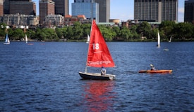 A sailboat with a prominent red sail and the number 18, featuring 'MIT' written on it, traverses a body of water with a person onboard. To the right, an orange kayak with a single individual paddling is visible. In the background, several other sailboats and the urban skyline of a city, marked by modern high-rise buildings and green trees, can be seen under a clear blue sky.