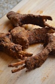 Several pieces of fried, breaded meat with visible bones are placed on a wooden cutting board. The meat appears crispy and well-seasoned.