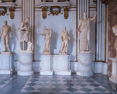A gallery room with several classical marble statues on ornate pedestals. The statues are arranged in a row, depicting various human figures in flowing robes and contrapposto stances. The room features luxurious decor with gold accents and intricate moldings. The floor has a diamond-patterned marble design.