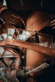 An intricate spiral staircase made of warm-toned wood and glass winds through an architectural space with a modern design. The structure features smooth curves and clean lines, with light filtering through vertical slats creating patterns on the floor. A person is in motion on the stairs, adding a touch of life to the serene, elegant setting.