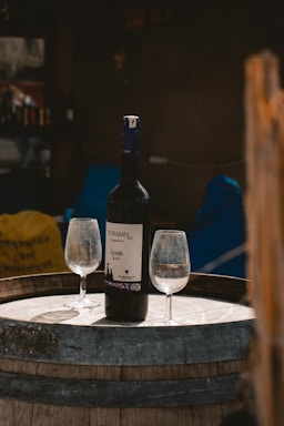 A bottle of Turasan Syrah 2020 wine stands on a wooden barrel, accompanied by two empty wine glasses. The setting appears to be outdoors, with a slightly rustic and casual ambiance.