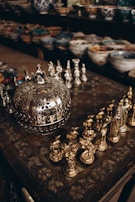 Intricately designed metallic chess pieces are arranged on an ornate table with a decorative pattern. In the background, shelves are filled with colorful ceramic bowls of various sizes and designs.