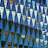 A geometric pattern of reflective triangular panels creates a mosaic effect. The panels display reflections of a cityscape, including buildings and windows, contrasting with the deep blue and turquoise colors of the glass. The angular shapes are arranged in a precise grid, giving an abstract and modern aesthetic.