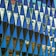 A geometric pattern of reflective triangular panels creates a mosaic effect. The panels display reflections of a cityscape, including buildings and windows, contrasting with the deep blue and turquoise colors of the glass. The angular shapes are arranged in a precise grid, giving an abstract and modern aesthetic.