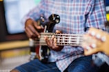 Fingers plucking the strings of a bass guitar, played by someone wearing a plaid shirt and denim shorts in a studio setting with blurred background.