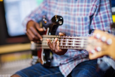 Fingers plucking the strings of a bass guitar, played by someone wearing a plaid shirt and denim shorts in a studio setting with blurred background.