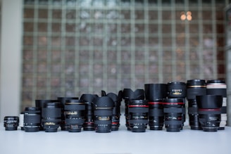 A collection of various camera lenses is arranged on a white surface. The lenses vary in size, shape, and brand, with some featuring distinguishable markings and labels. The background consists of a wall made of glass blocks, adding a patterned, semi-transparent backdrop to the scene.