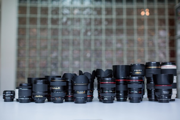A collection of various camera lenses is arranged on a white surface. The lenses vary in size, shape, and brand, with some featuring distinguishable markings and labels. The background consists of a wall made of glass blocks, adding a patterned, semi-transparent backdrop to the scene.