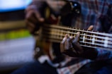 A person playing an electric bass guitar, with close-up focus on the musician's hands and guitar strings. The background is blurred, highlighting the instrument and fingers.
