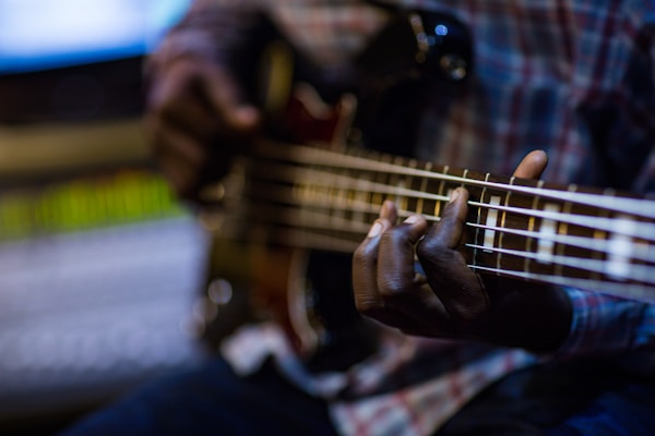 A person playing an electric bass guitar, with close-up focus on the musician's hands and guitar strings. The background is blurred, highlighting the instrument and fingers.