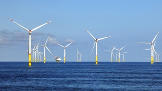 Several wind turbines are positioned in the ocean, creating an offshore wind farm. The turbines are evenly distributed across the water, which is calm and reflects a shade of blue. The sky above is mostly clear with a few clouds.