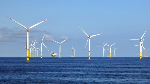 Several wind turbines are positioned in the ocean, creating an offshore wind farm. The turbines are evenly distributed across the water, which is calm and reflects a shade of blue. The sky above is mostly clear with a few clouds.