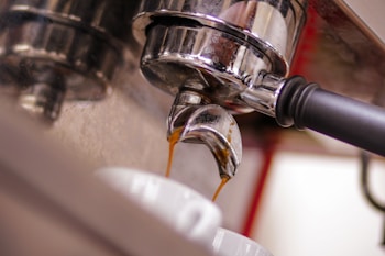 Coffee is being extracted from an espresso machine with a metal portafilter into a white cup. The focus is on the portafilter and the stream of rich, dark coffee, highlighting the shiny chrome finish and the smooth flow of the espresso.