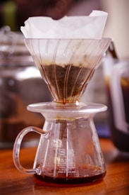 A glass pour-over coffee maker with a paper filter placed on top sits on a wooden surface. The filter contains ground coffee, and freshly brewed coffee is dripping into the glass carafe below. The background is slightly blurred, highlighting the coffee-making process.