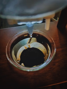 A close-up view of coffee dripping into a filter set within a glass coffee maker. The coffee grounds are dark and finely ground, and the setup is placed on a wooden surface. The process of brewing coffee manually is shown with the focus on the drip and filter.