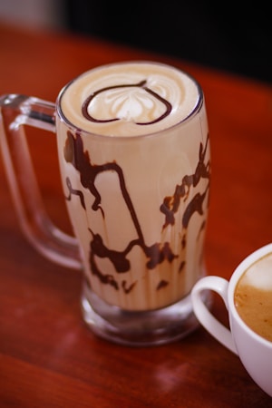 A glass mug filled with a creamy iced coffee topped with a swirl of whipped cream and chocolate drizzle. The mug is accented with artistic chocolate patterns on the inside. Next to it, a white cup holds a frothy cappuccino.