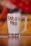 A clear glass mug filled with a creamy beverage has 'Barista Pro' written in chocolate syrup on its surface. The mug is set against a blurred background with warm tones of red, orange, and yellow, creating a cozy atmosphere.