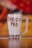 A clear glass mug filled with a creamy beverage has 'Barista Pro' written in chocolate syrup on its surface. The mug is set against a blurred background with warm tones of red, orange, and yellow, creating a cozy atmosphere.