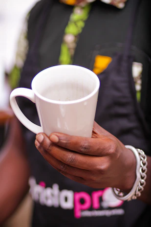 A person holding a white ceramic mug with their right hand visible, wearing a bracelet and a black apron with colorful patterns underneath.