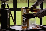 A person wearing a patterned shirt is pouring a glass of whiskey or a similar beverage from a bottle. The scene includes a wooden table or tray with a box labeled 'Lagavulin 16', a couple of glasses, one with liquid and the other empty, a glass decanter, and a wristwatch visible on the person's wrist. The background shows a green outdoor area through a window.